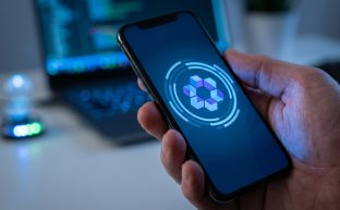 Modern bir teknoloji ortamında, ekranda soyut bir güvenlik kodu gösteren bir kimlik doğrulama uygulamasının açık olduğu bir akıllı telefonu tutan bir elin yakın çekim fotoğrafı. Arka plan, hesap girişi ve dijital güvenlik konseptini ima eden bulanık bir dizüstü bilgisayardan oluşuyor.
