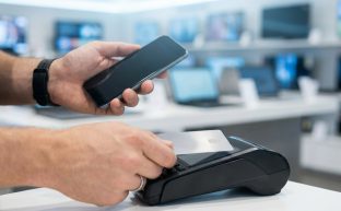 Modern bir teknoloji mağazasının bulanık arka planında, bir kişinin elinde akıllı telefon tutarken diğer eliyle jenerik bir kredi kartını POS cihazına yaklaştırarak temassız ödeme yaptığı anın yakın çekim fotoğrafı. Görselde hiçbir yazı veya logo bulunmamaktadır.