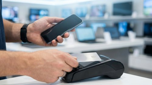 Modern bir teknoloji mağazasının bulanık arka planında, bir kişinin elinde akıllı telefon tutarken diğer eliyle jenerik bir kredi kartını POS cihazına yaklaştırarak temassız ödeme yaptığı anın yakın çekim fotoğrafı. Görselde hiçbir yazı veya logo bulunmamaktadır.